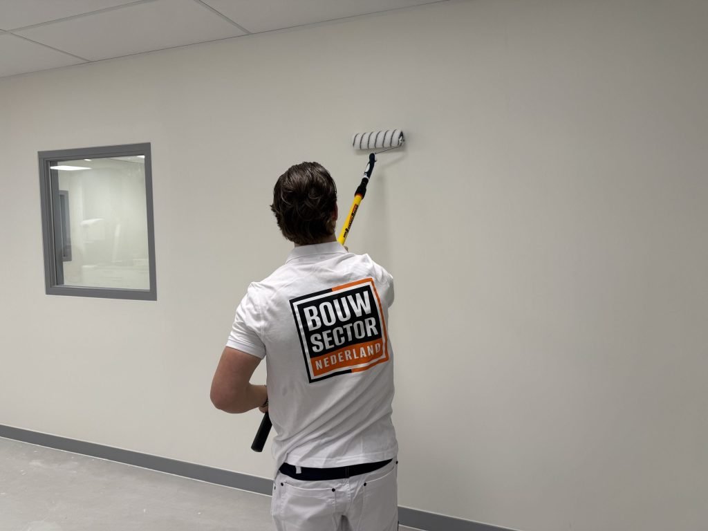 Schilder van Bouwsector Nederland rolt een gladde wand tot tegen het plafond, naast een binnenraam